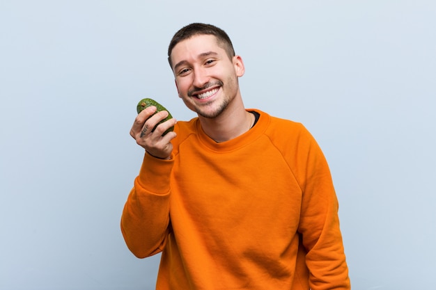 Jovem homem caucasiano segurando um abacate feliz, sorridente e alegre.