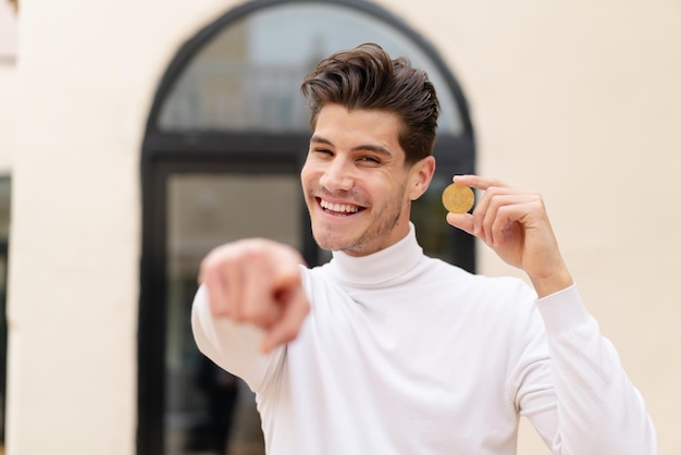 Jovem homem caucasiano segurando um Bitcoin ao ar livre aponta o dedo para você com uma expressão confiante