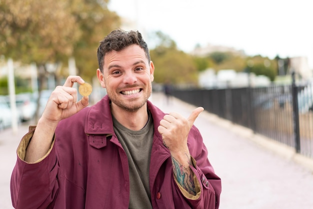 Jovem homem caucasiano segurando um Bitcoin ao ar livre apontando para o lado para apresentar um produto