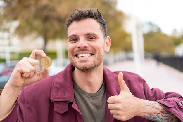 Jovem homem caucasiano segurando um Bitcoin ao ar livre com polegares para cima porque algo bom aconteceu