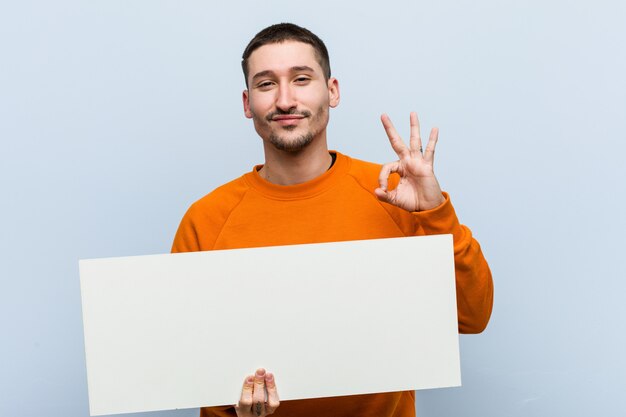 Jovem homem caucasiano segurando um cartaz alegre e confiante mostrando okey gesto.