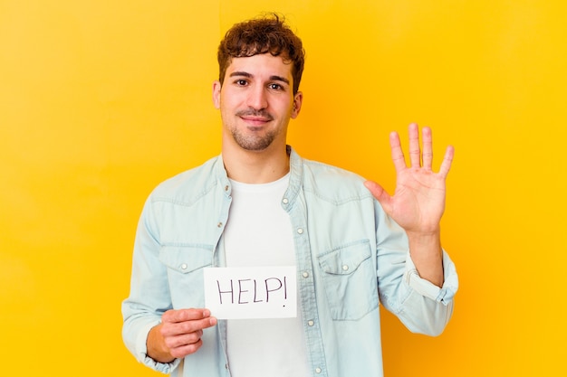 Jovem homem caucasiano segurando um cartaz de ajuda isolado sorrindo alegre mostrando o número cinco com os dedos.
