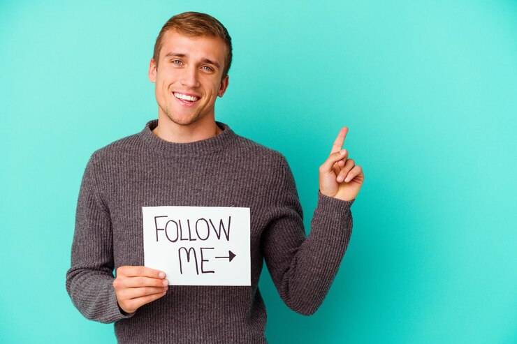 Foto jovem homem caucasiano segurando um cartaz de siga-me isolado no azul, sorrindo alegremente, apontando com o dedo indicador para longe.