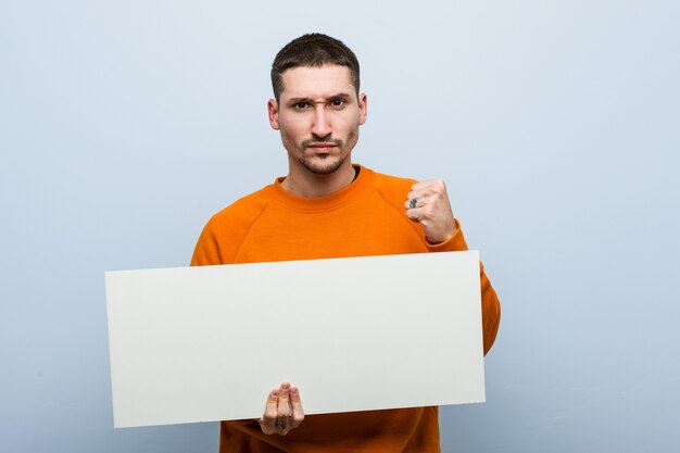 Jovem homem caucasiano segurando um cartaz mostrando o punho, expressão facial agressiva.