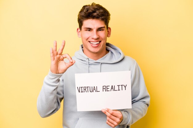 Jovem homem caucasiano segurando um cartaz virtual isolado em fundo amarelo alegre e confiante, mostrando o gesto okey