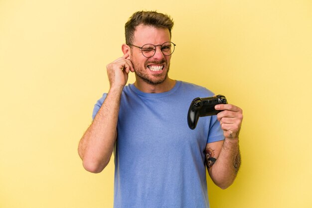 Jovem homem caucasiano, segurando um controlador de jogo isolado em fundo amarelo, cobrindo os ouvidos com as mãos.