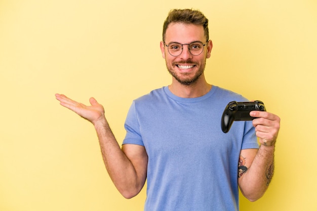 Jovem homem caucasiano segurando um controlador de jogo isolado em fundo amarelo, mostrando um espaço de cópia na palma da mão e segurando a outra mão na cintura.