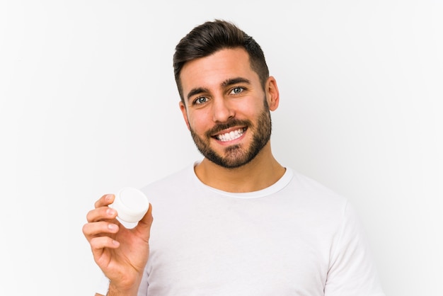 Jovem homem caucasiano segurando um creme hidratante isolado feliz, sorridente e alegre.