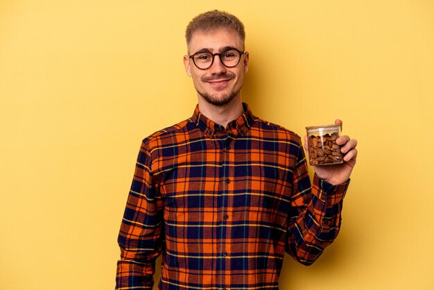 Jovem homem caucasiano segurando um frasco de amêndoa isolado em fundo amarelo feliz, sorridente e alegre.