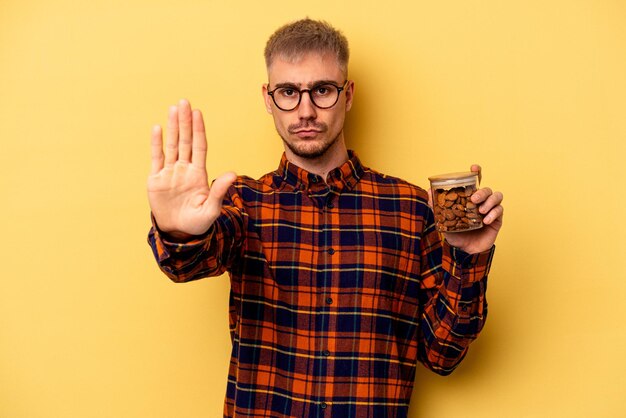 Jovem homem caucasiano segurando um frasco de amêndoa isolado no fundo amarelo em pé com a mão estendida, mostrando o sinal de pare, impedindo você.