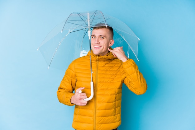 Jovem homem caucasiano segurando um guarda-chuva, mostrando um gesto de chamada de telefone móvel com os dedos.