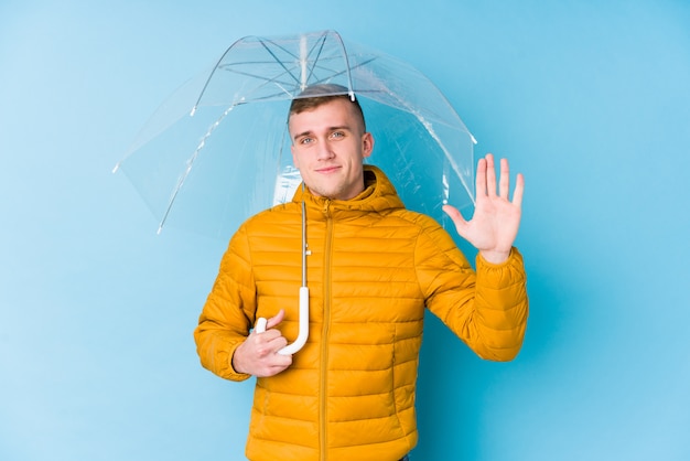 Foto jovem homem caucasiano segurando um guarda-chuva sorrindo alegre mostrando o número cinco com os dedos.