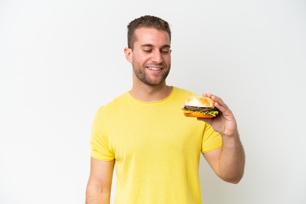 Jovem homem caucasiano segurando um hambúrguer isolado no fundo branco com expressão feliz