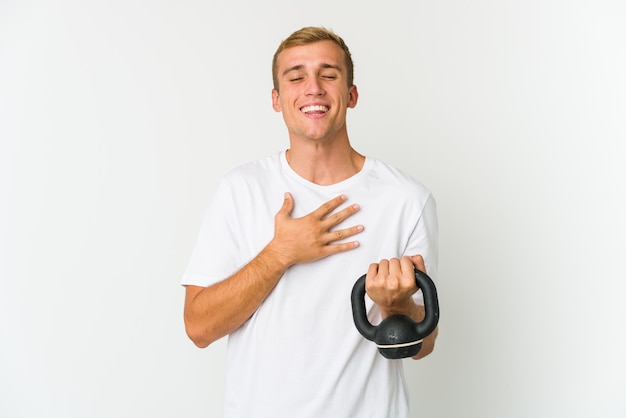Jovem homem caucasiano segurando um kettlebell isolado na parede branca, ri alto, mantendo a mão no peito.