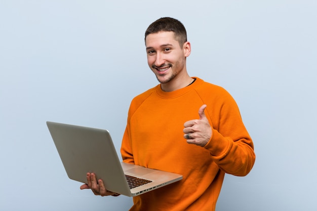 Jovem homem caucasiano segurando um laptop sorrindo e levantando o polegar para cima