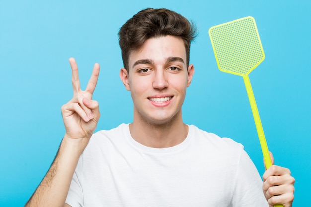 Jovem homem caucasiano segurando um mata-moscas, mostrando sinal de vitória e sorrindo amplamente.