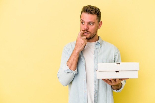 Jovem homem caucasiano segurando um pizzas isoladas em fundo amarelo relaxado pensando em algo olhando para um espaço de cópia.