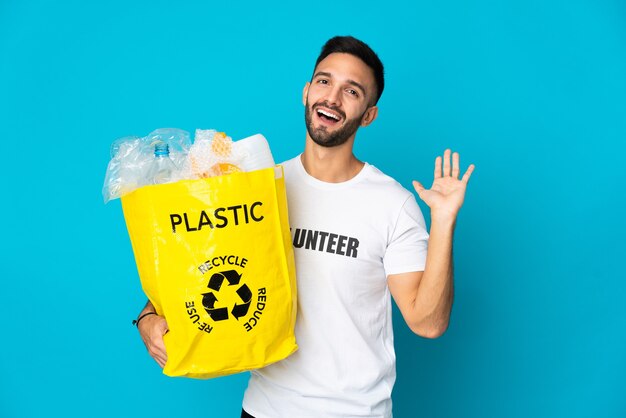 Jovem homem caucasiano segurando um saco cheio de garrafas plásticas para reciclar isolado em um fundo azul, saudando com a mão com expressão feliz