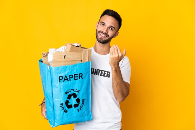 Jovem homem caucasiano segurando um saco de reciclagem cheio de papel para reciclar isolado na parede branca, convidando a vir com a mão. Feliz que você veio