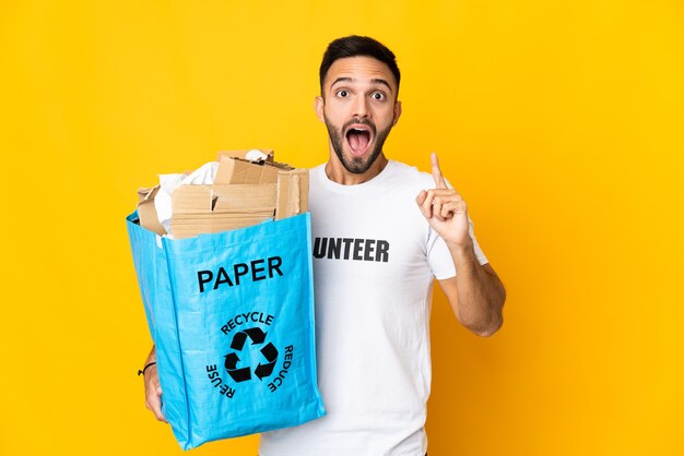 Jovem homem caucasiano segurando um saco de reciclagem cheio de papel para reciclar isolado no fundo branco com a intenção de perceber a solução enquanto levanta um dedo