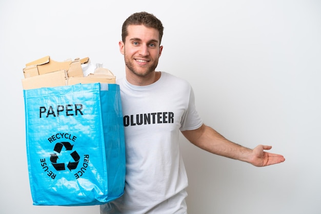Jovem homem caucasiano segurando um saco de reciclagem cheio de papel para reciclar isolado no fundo branco estendendo as mãos para o lado para convidar para vir