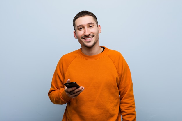 Jovem homem caucasiano segurando um telefone feliz, sorridente e alegre.