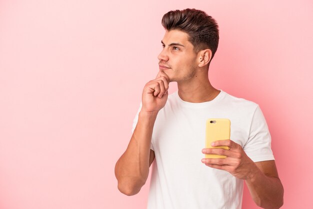 Jovem homem caucasiano segurando um telefone móvel isolado no fundo rosa, olhando de soslaio com expressão duvidosa e cética.