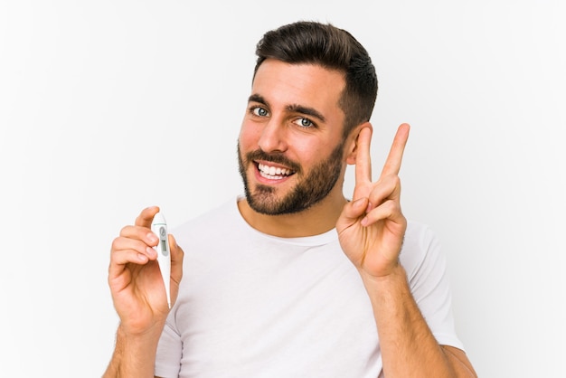 Jovem homem caucasiano segurando um termômetro isolado Jovem homem caucasiano segurando um sinal de vitória mostrando e sorrindo amplamente.