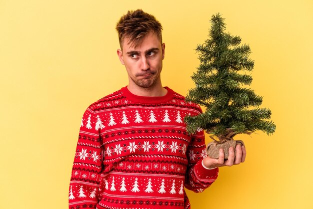 Jovem homem caucasiano segurando uma árvore de Natal isolada em um fundo amarelo confuso, sente-se em dúvida e inseguro.