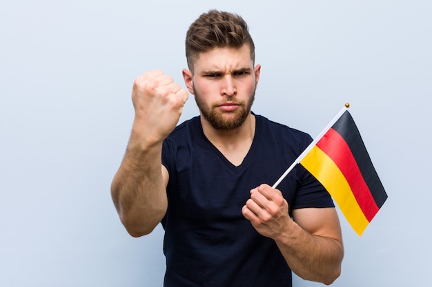 Jovem homem caucasiano segurando uma bandeira da Alemanha, mostrando o punho para a câmera, expressão facial agressiva.