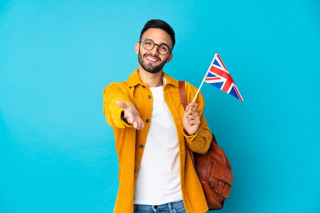 Jovem homem caucasiano segurando uma bandeira do Reino Unido isolada em fundo amarelo apertando as mãos para fechar um bom negócio