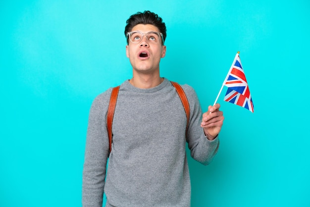 Jovem homem caucasiano segurando uma bandeira do Reino Unido isolada em fundo azul olhando para cima e com expressão de surpresa