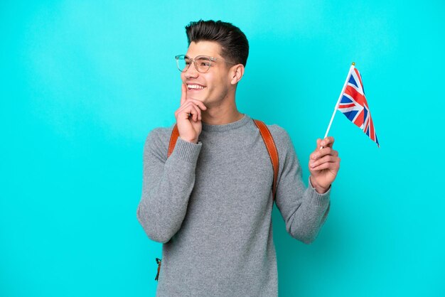 Jovem homem caucasiano segurando uma bandeira do Reino Unido isolada em fundo azul pensando em uma ideia enquanto olha para cima