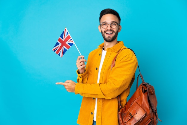 Jovem homem caucasiano segurando uma bandeira do Reino Unido isolada em um fundo amarelo apontando o dedo para o lado