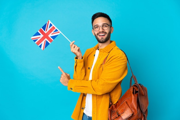 Jovem homem caucasiano segurando uma bandeira do Reino Unido isolada em um fundo amarelo apontando para trás