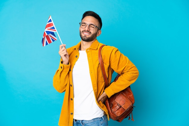 Jovem homem caucasiano segurando uma bandeira do Reino Unido isolada em um fundo amarelo, sofrendo de dor nas costas por ter feito um esforço