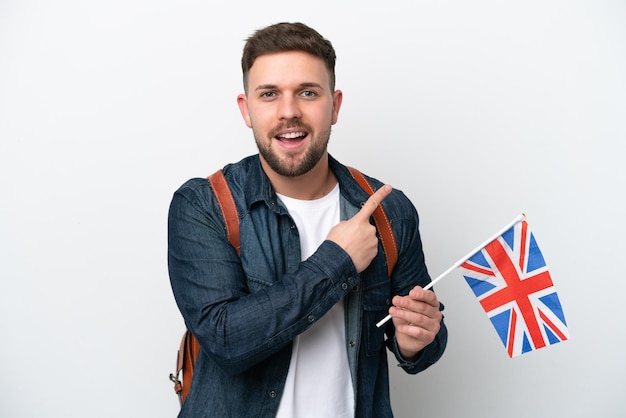 Jovem homem caucasiano segurando uma bandeira do Reino Unido isolada no fundo branco, apontando para o lado para apresentar um produto