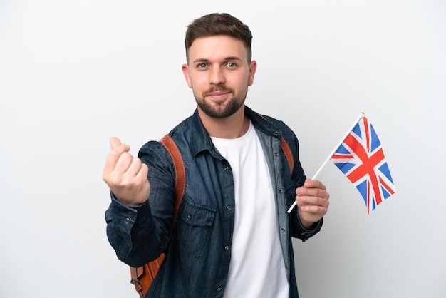 Jovem homem caucasiano segurando uma bandeira do Reino Unido isolada no fundo branco, fazendo gesto de dinheiro