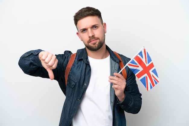 Jovem homem caucasiano segurando uma bandeira do Reino Unido isolada no fundo branco, mostrando o polegar para baixo com expressão negativa