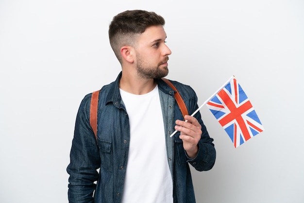 Jovem homem caucasiano segurando uma bandeira do Reino Unido isolada no fundo branco, olhando para o lado