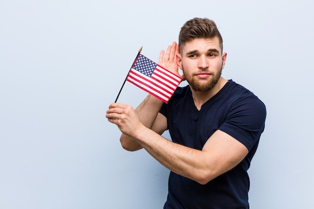 Jovem homem caucasiano segurando uma bandeira dos Estados Unidos, tentando ouvir uma fofoca.