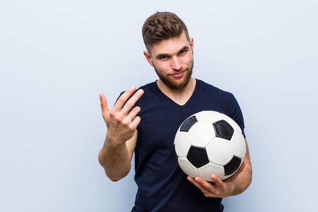 Jovem homem caucasiano segurando uma bola de futebol, apontando com o dedo para você, como se estivesse convidando para se aproximar.