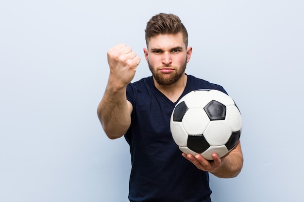 Jovem homem caucasiano segurando uma bola de futebol, mostrando o punho para a câmera, expressão facial agressiva.