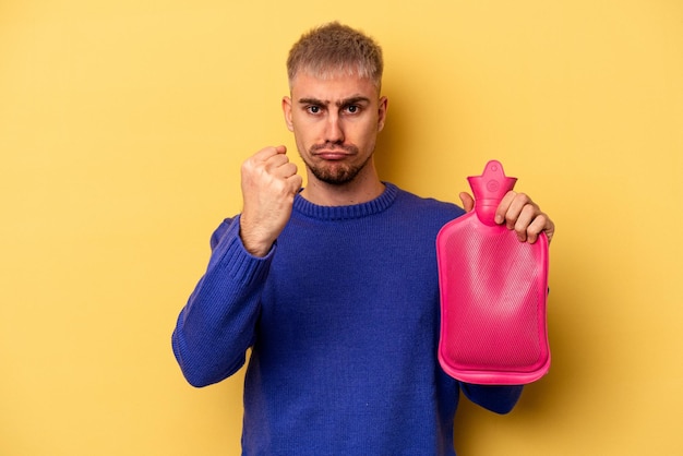 Jovem homem caucasiano segurando uma bolsa de água isolada em fundo amarelo, mostrando o punho para a câmera, expressão facial agressiva.