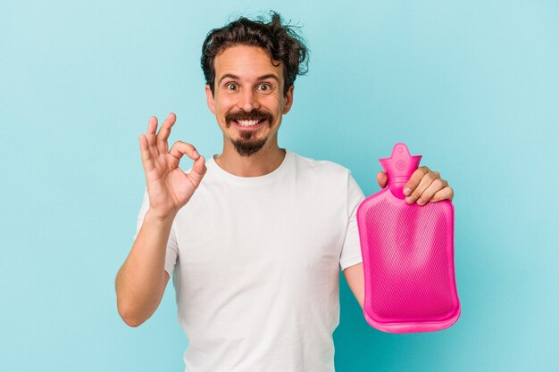 Jovem homem caucasiano segurando uma bolsa de água isolada no fundo azul alegre e confiante mostrando o gesto de ok.