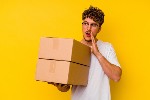 Jovem homem caucasiano segurando uma caixa de papelão isolada na parede amarela está contando uma notícia secreta da frenagem e olhando para o lado