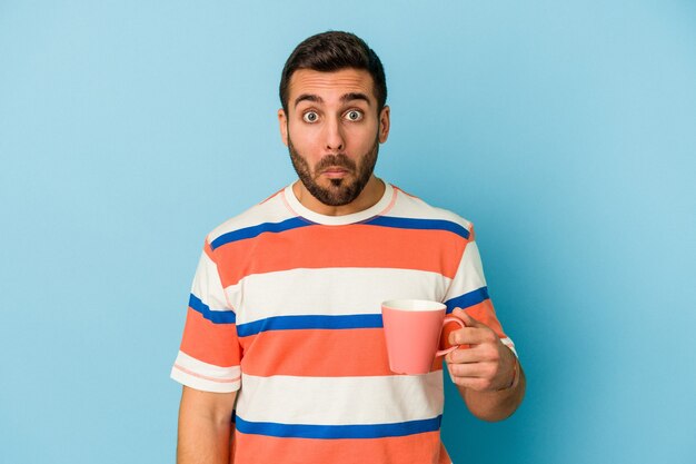 Jovem homem caucasiano segurando uma caneca isolada sobre fundo azul encolhe os ombros e abre os olhos confusos.