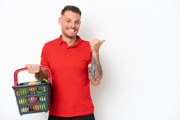 Jovem homem caucasiano segurando uma cesta de compras cheia de comida isolada no fundo branco apontando para o lado para apresentar um produto