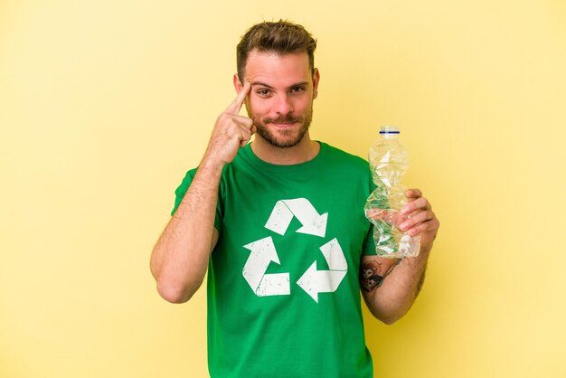 Jovem homem caucasiano segurando uma garrafa de plástico para reciclar isolado em fundo amarelo, apontando o templo com o dedo, pensando, focado em uma tarefa.