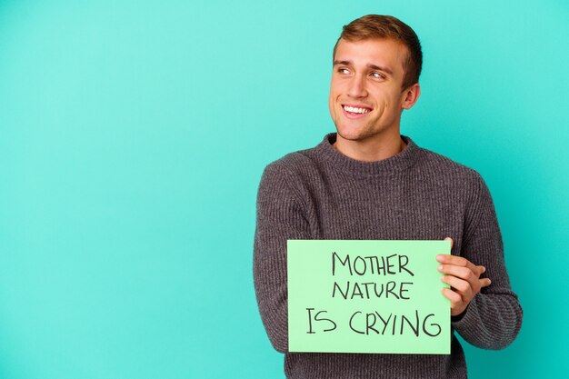 Jovem homem caucasiano segurando uma mãe natureza está chorando cartaz isolado no azul, sorrindo confiante com os braços cruzados.